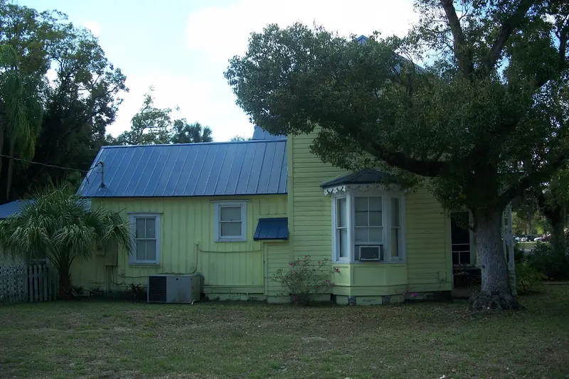 Small yellow wood-sided cottage in Florida with a blue metal roof, half of it under the spreading shade of a broad oak, a window unit and condenser along the side wall