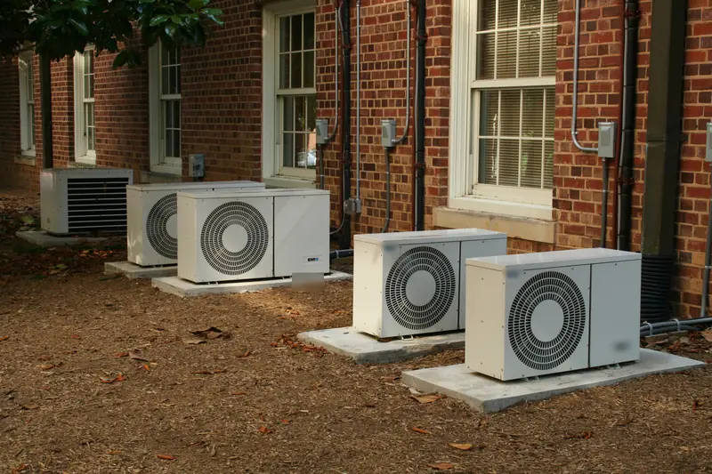 Four white outdoor heat pump units lined up on concrete pads against a red brick wall